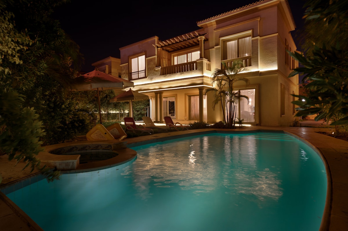 An elegant mansion is illuminated by soft lighting at night. The crystal-clear pool reflects the glow, surrounded by lounge chairs and lush greenery. A Jacuzzi sits nearby, enhancing the inviting outdoor space that seamlessly connects to the house’s large windows and warm ambiance.
