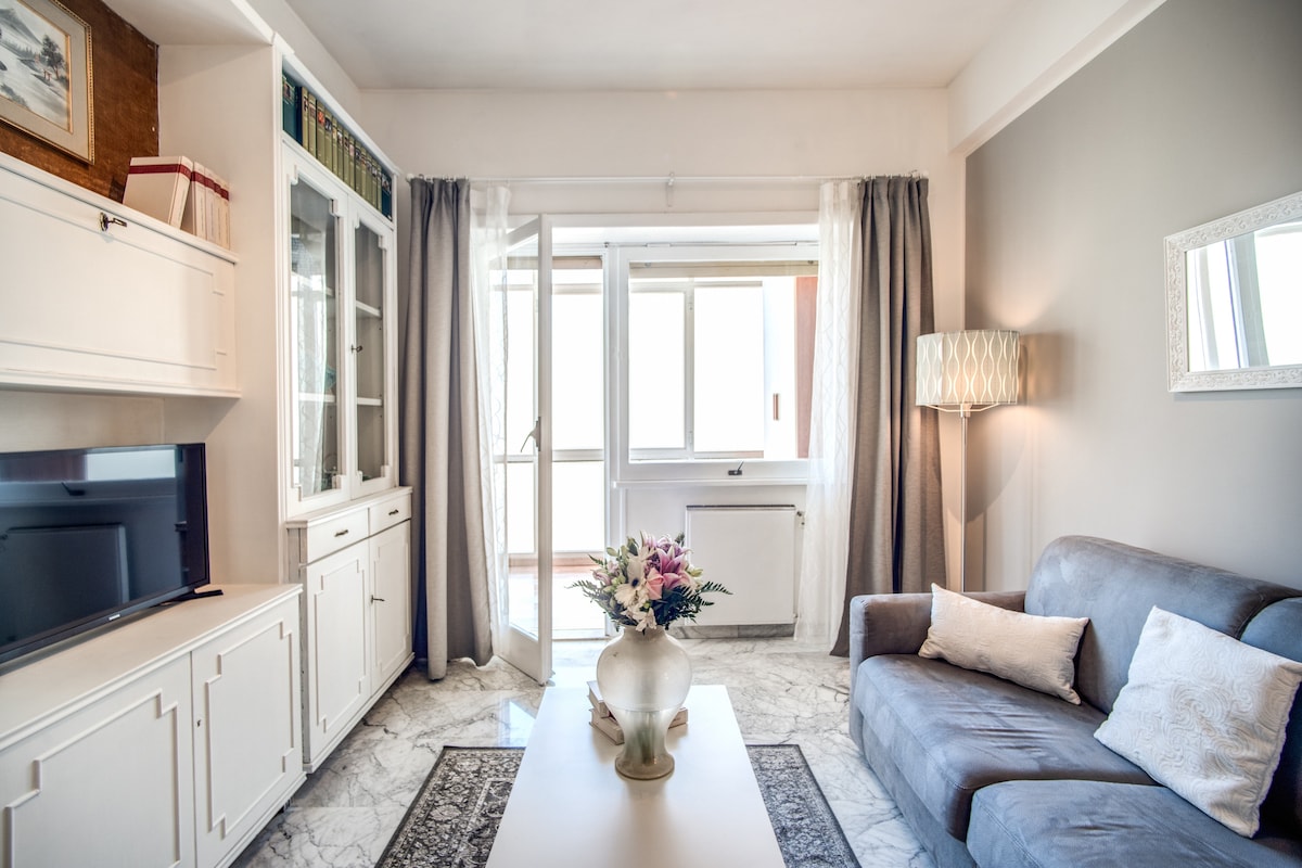 A bright living room features a comfortable gray sofa facing a wall unit with a television. Natural light enters through sliding glass doors that lead to the outside. A decorative vase with flowers rests on a central table, complemented by soft curtains.