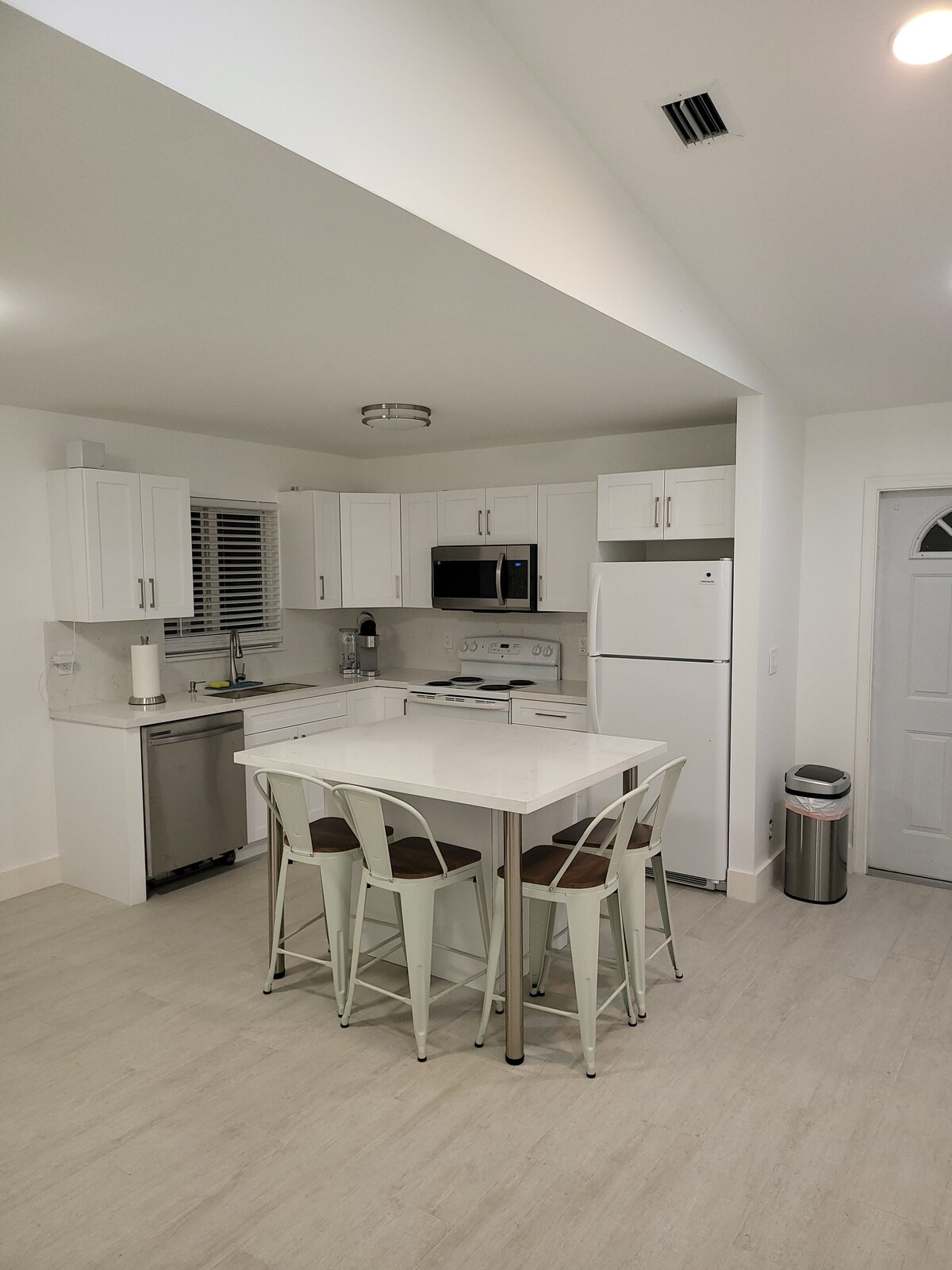 A modern kitchen is showcased, featuring white cabinetry and stainless-steel appliances. A circular table with four chairs occupies the space, complemented by a large window allowing natural light. The neutral-toned floor adds to the overall brightness and airy feel of the area.