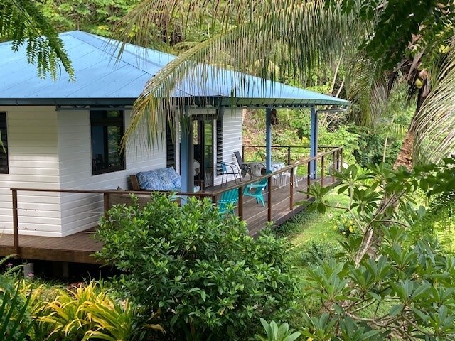 A single-story house with a blue roof is framed by lush greenery. A covered porch features outdoor seating and overlooks the landscaped garden, providing a serene view. The structure is surrounded by vibrant plants and trees, emphasizing a peaceful natural environment.
