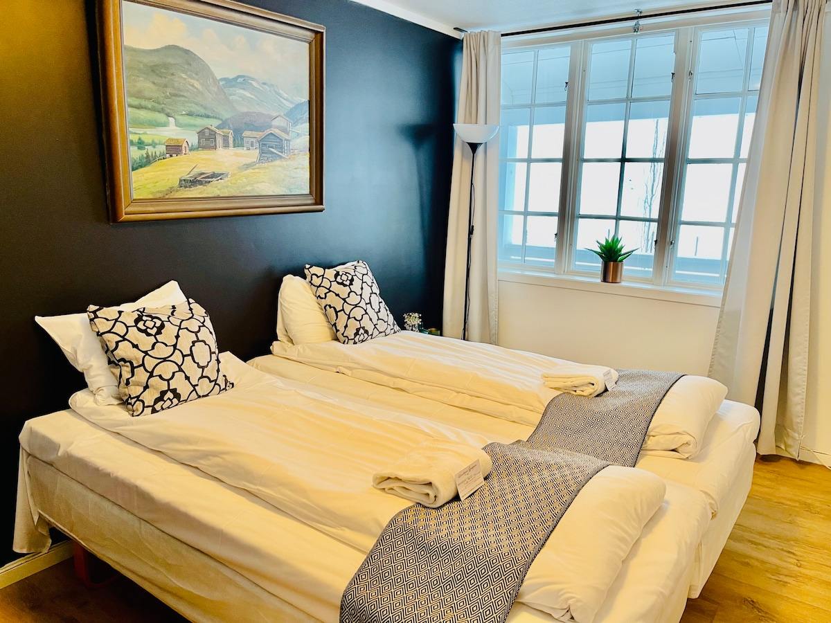 A well-lit sleeping area features two spacious beds dressed in crisp white linens. Decorative throw pillows add comfort. A landscape painting hangs above the beds, while a potted plant and light-filtering window treatments enhance the room's welcoming feel.