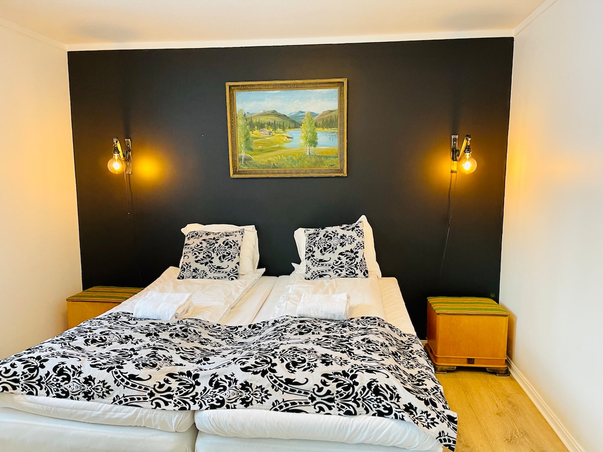 A sleeping area features two spacious beds adorned with black and white patterned bedding. Decorative pillows complement the design. Soft lighting is provided by two fixtures attached to a dark accent wall, while a framed landscape painting adds a touch of character to the room.