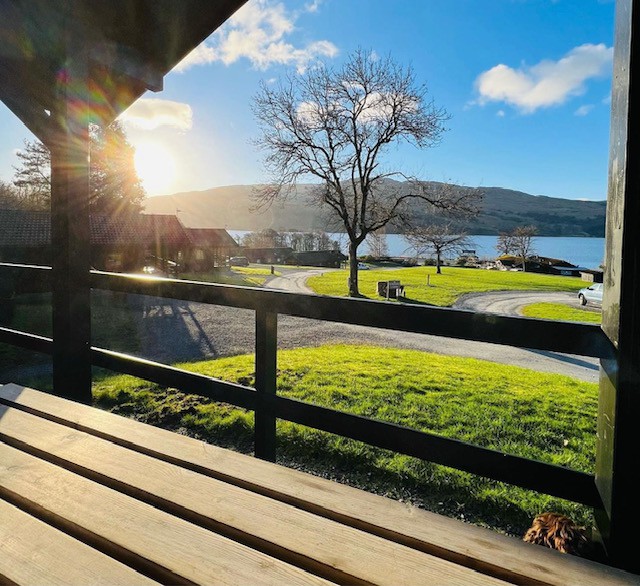 A view from the lodge's porch captures the early morning sun illuminating the landscape. A tree stands in the foreground, with Loch Tay visible in the background, framed by rolling hills and scattered buildings along the shore.