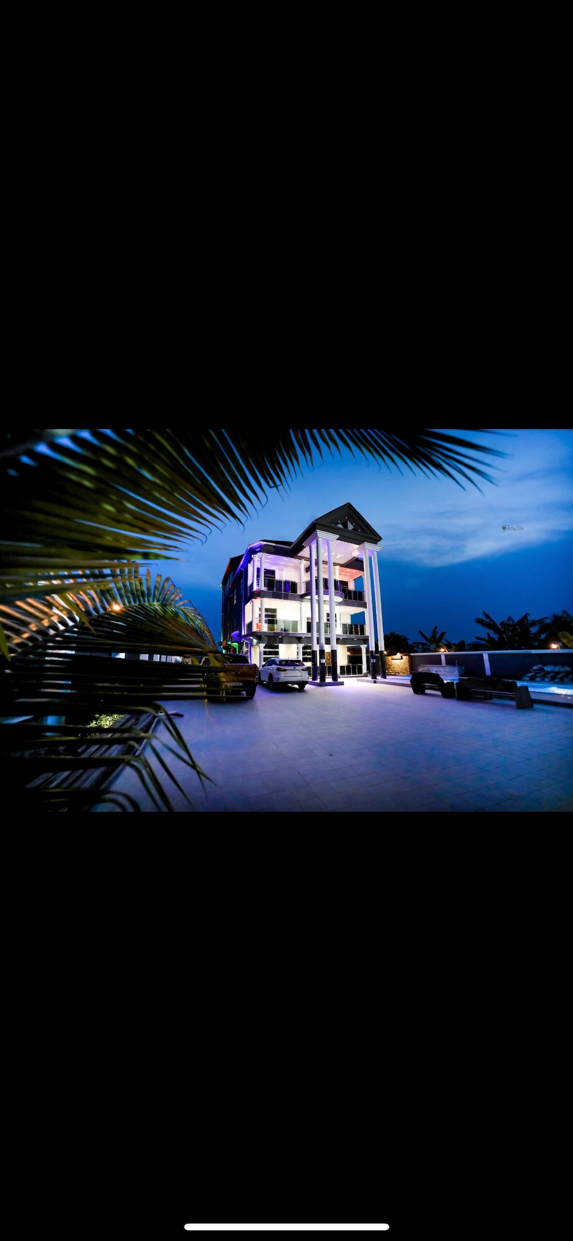 A modern three-story building is showcased against a twilight sky, highlighted by exterior lighting. Palm fronds frame the image, while vehicles are visible parked in front of the entrance. The clean lines of the architecture contribute to a contemporary aesthetic.