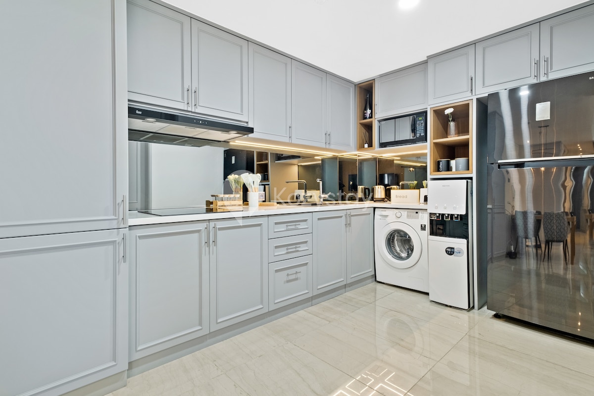 A modern kitchen features sleek gray cabinetry and polished countertop lighting. Equipped with essential appliances, including a refrigerator, a microwave, and an induction stove. An in-suite washer is also included, enhancing convenience for guests.