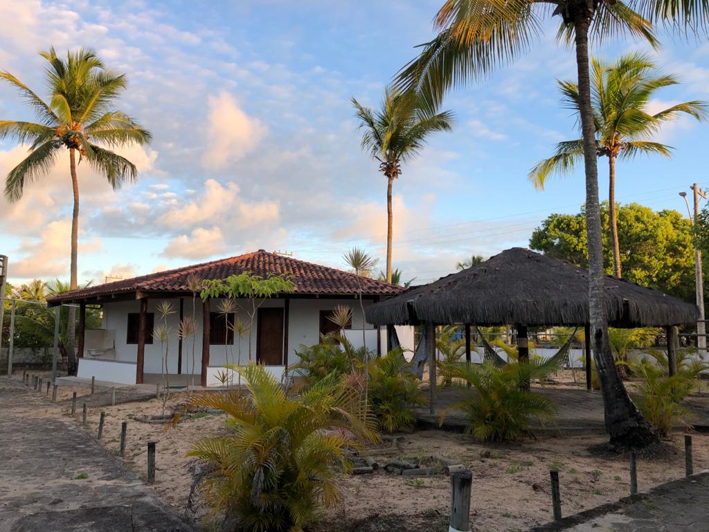 A single-story building with a tiled roof is situated beside a thatched gazebo. Lush greenery, including palm trees and small shrubs, surrounds the property. The sandy area suggests a relaxed outdoor space, ideal for gatherings or leisure under the sun.