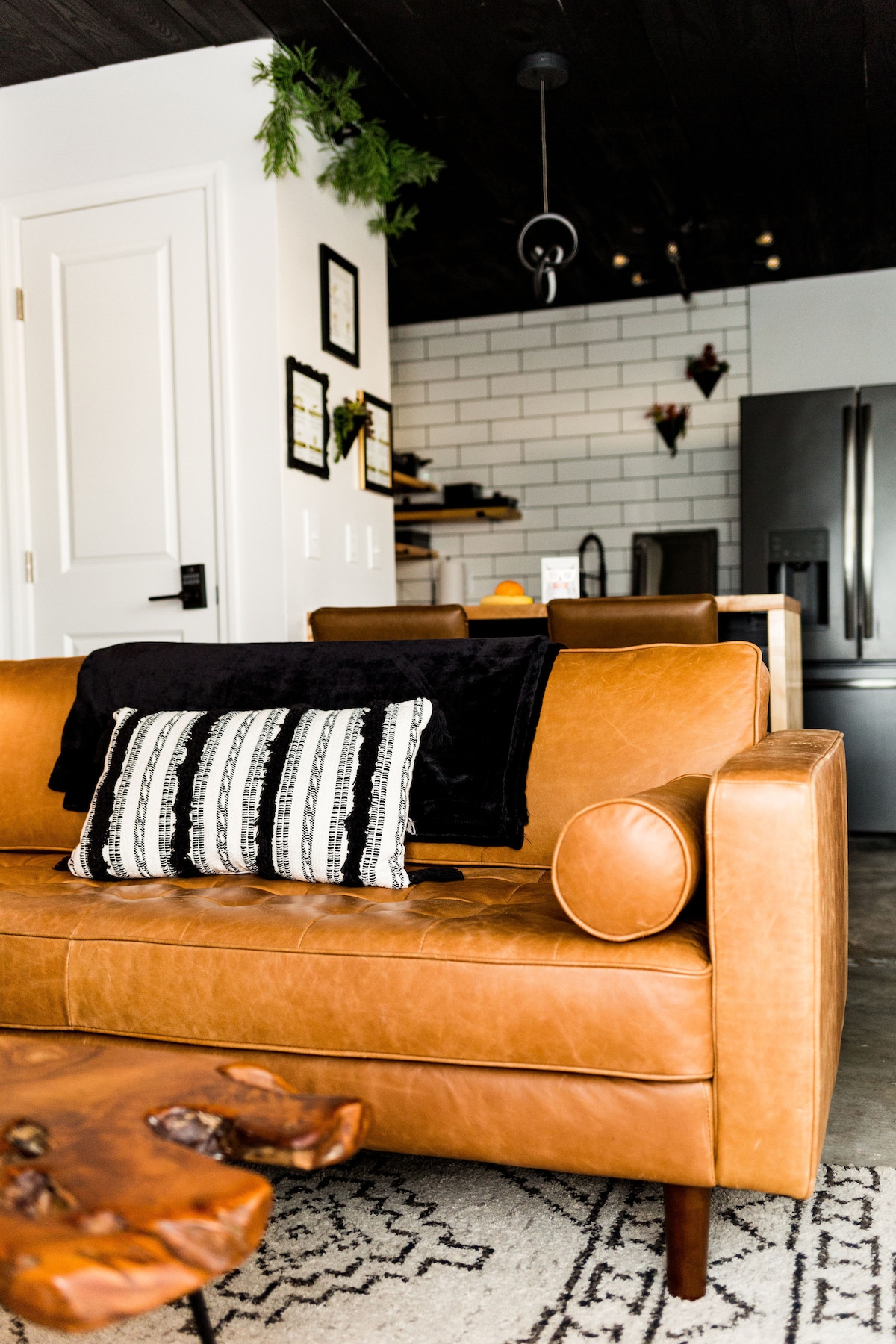 A cozy leather couch is positioned in the living room, featuring decorative pillows. A unique wooden coffee table adds character to the space. The kitchen is visible in the background, showcasing modern appliances and a minimalist design.