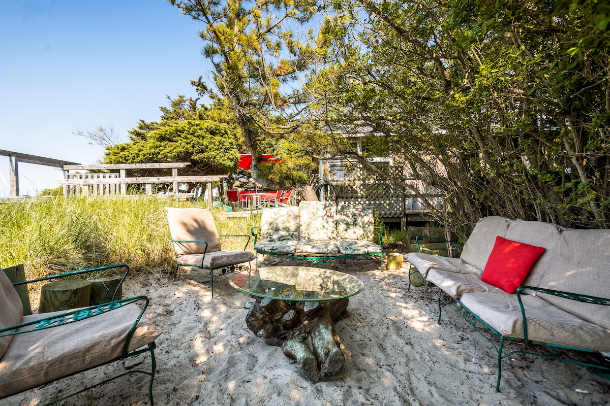 A sandy outdoor sitting area is surrounded by lush greenery. A low glass table is set on a natural wood base, surrounded by four comfortable chairs with light fabric cushions. Soft red accents enhance the relaxing atmosphere, inviting guests to unwind in nature.