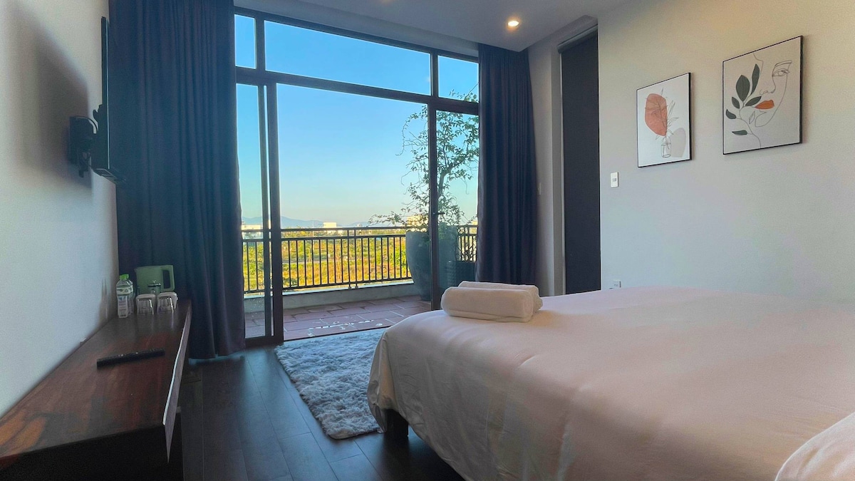 A well-lit bedroom features a spacious bed with a rolled towel positioned neatly on top. Large glass doors open to a balcony with a scenic view, allowing natural light to flow in. Decor includes minimalist wall art and a dark curtain complementing the wooden floor.