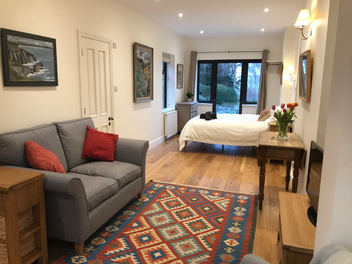 An open-plan studio features a spacious bed and a cozy seating area with a grey sofa and wooden side table. An area rug adds color beneath the furniture. Large windows invite natural light and offer views of the surrounding garden.