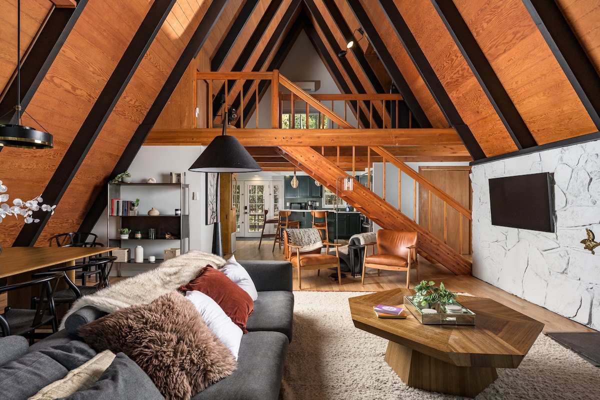 A warm living space is highlighted by high, contrasting wooden beams and a stone accent wall. A spacious gray sofa adorned with throw pillows faces a wooden coffee table. Cozy seating and a dining area are visible, enhancing the inviting atmosphere of the A-frame interior.