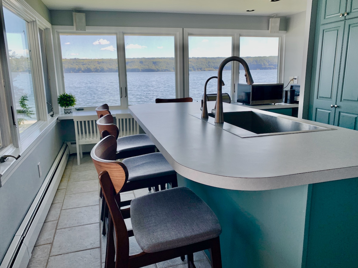 The kitchen features a spacious island with seating for five and sleek bar stools. Large windows provide views of the lake, allowing natural light to flood the area. A modern faucet above the sink complements the understated cabinetry and counter space.