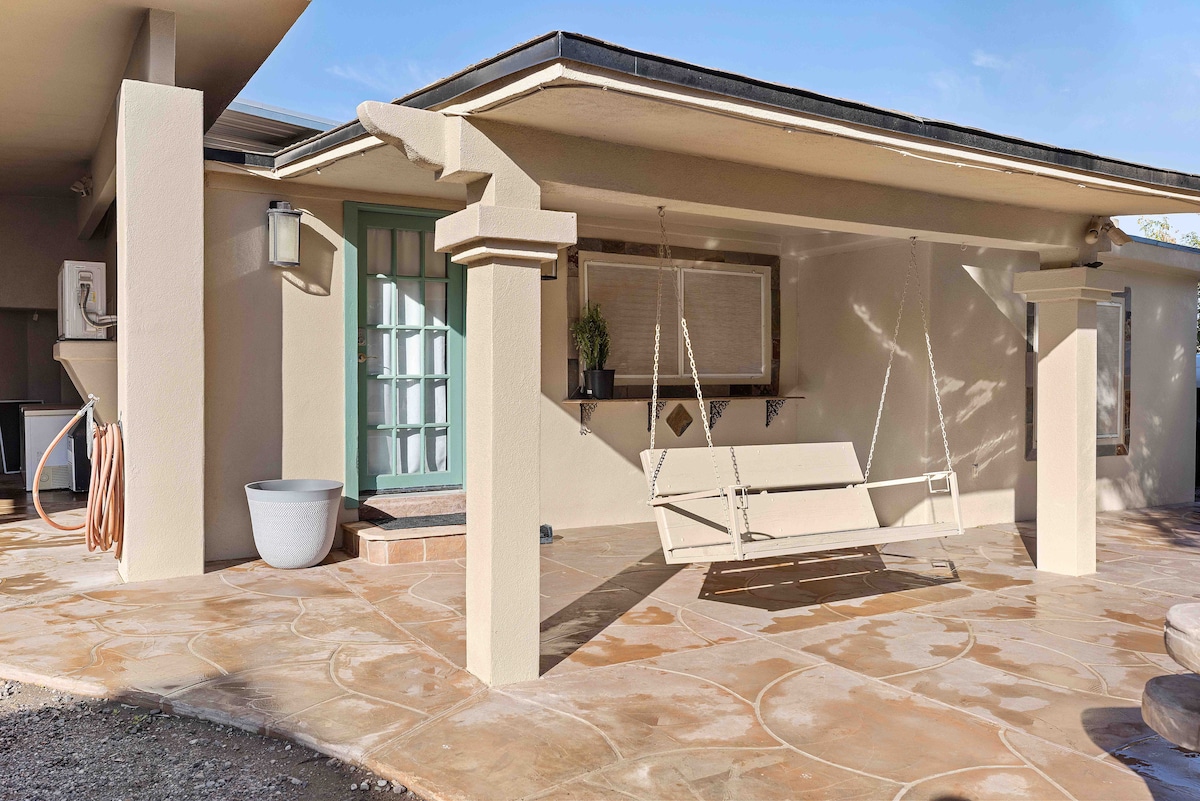 A charming porch features a swing suspended from the ceiling, offering a relaxed seating option. The textured flooring complements the neutral exterior walls, while a light blue door adds a touch of color. A small potted plant decorates the wall, enhancing the welcoming environment.