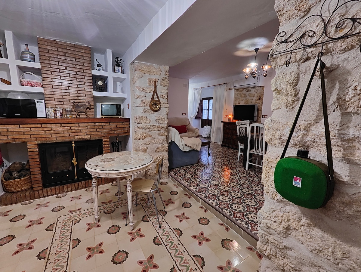 A rustic dining area features a round table surrounded by white chairs, set against a backdrop of patterned flooring. A cozy seating area is visible, alongside a traditional brick fireplace and open shelving displaying decorative items. Soft lighting is provided by overhead fixtures.
