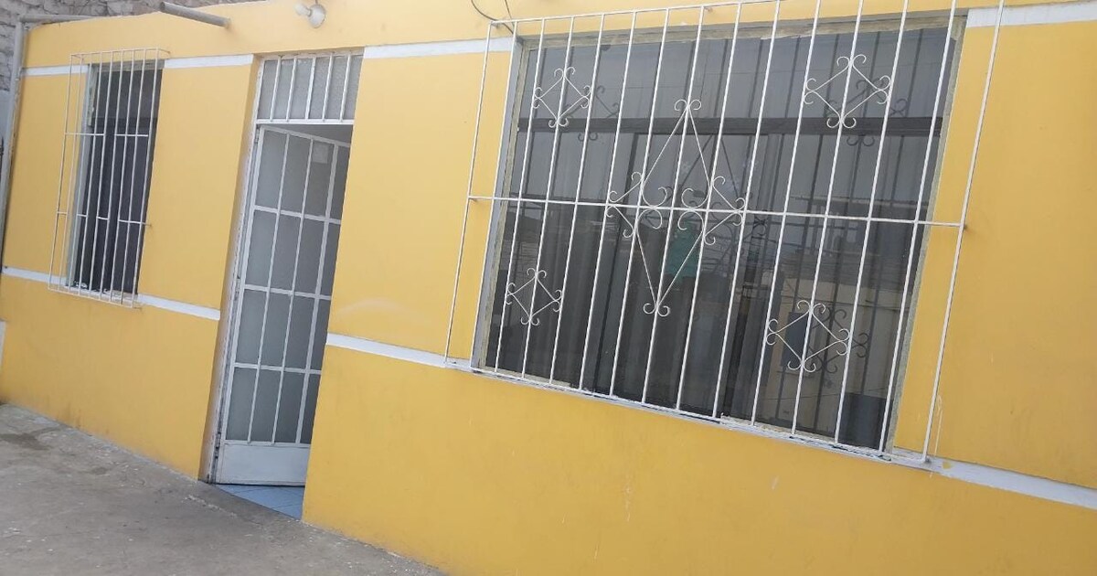 A cheerful yellow exterior is shown, featuring a glass door and several large, barred windows that allow natural light indoors. The walls are complemented by a simple concrete walkway, creating a welcoming entrance.