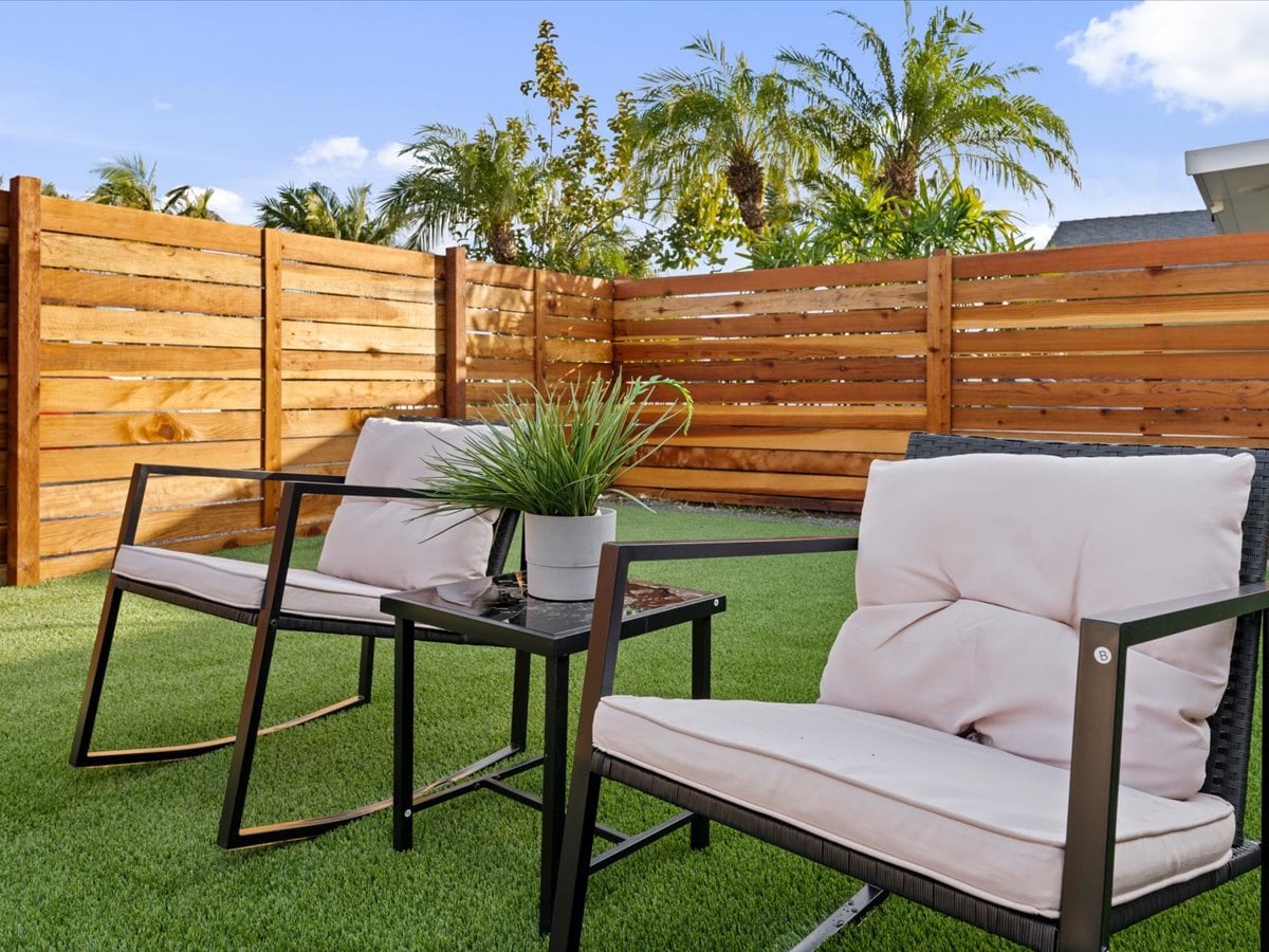 Two comfortable chairs are positioned around a small table on a private lawn. The space is bordered by wooden fencing and features lush greenery, providing a serene spot for relaxation.
