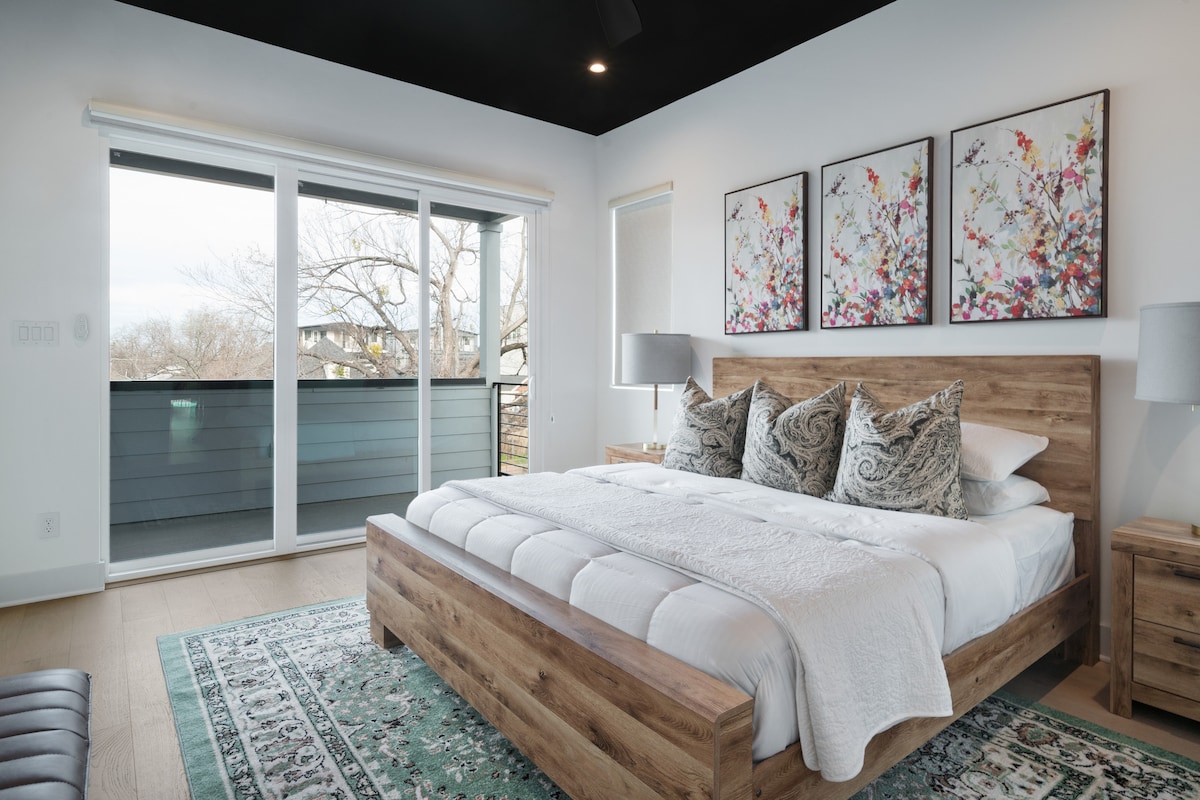 A well-appointed bedroom features a wooden bed frame with a spacious mattress and decorative throw pillows. Large sliding glass doors open to a balcony, allowing natural light to illuminate the space. Floral artwork adorns the walls, providing a soothing atmosphere.