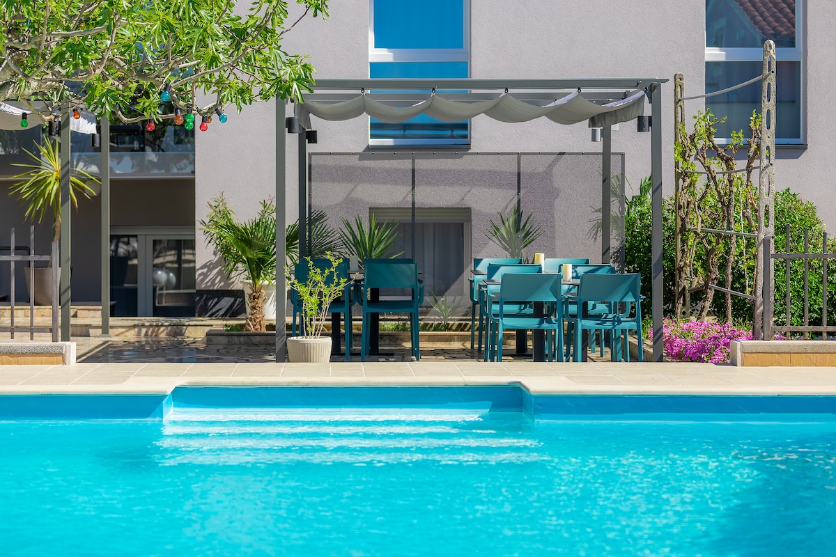 A spacious terrace offers an outdoor dining area shaded by a pergola, featuring a large table surrounded by blue chairs. Lush greenery and flowering plants frame the area, while a sparkling swimming pool is visible in the foreground with gentle steps leading into the water.