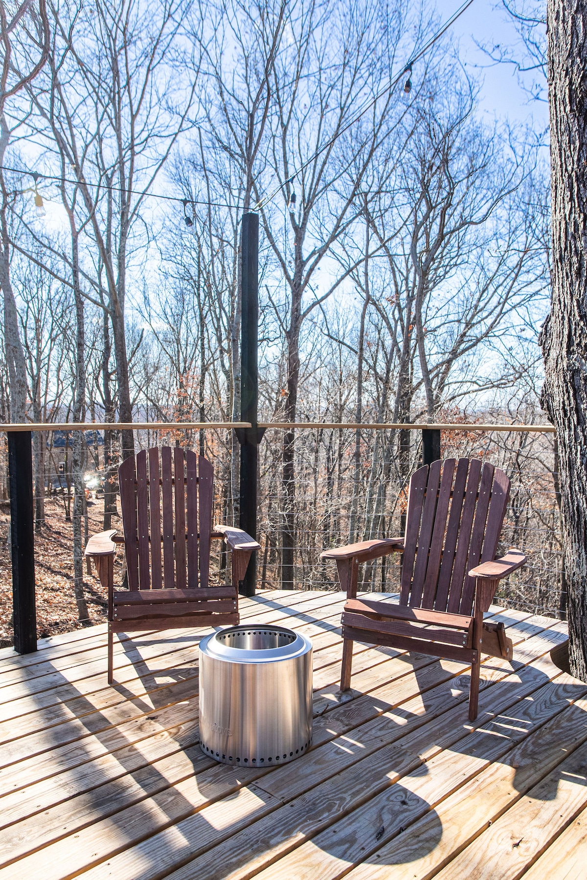 A spacious wooden deck features two classic Adirondack chairs facing the treetops. A stainless steel fire pit sits in the center, surrounded by natural wood flooring. The surrounding trees stand bare, allowing for scenic views of the forest.
