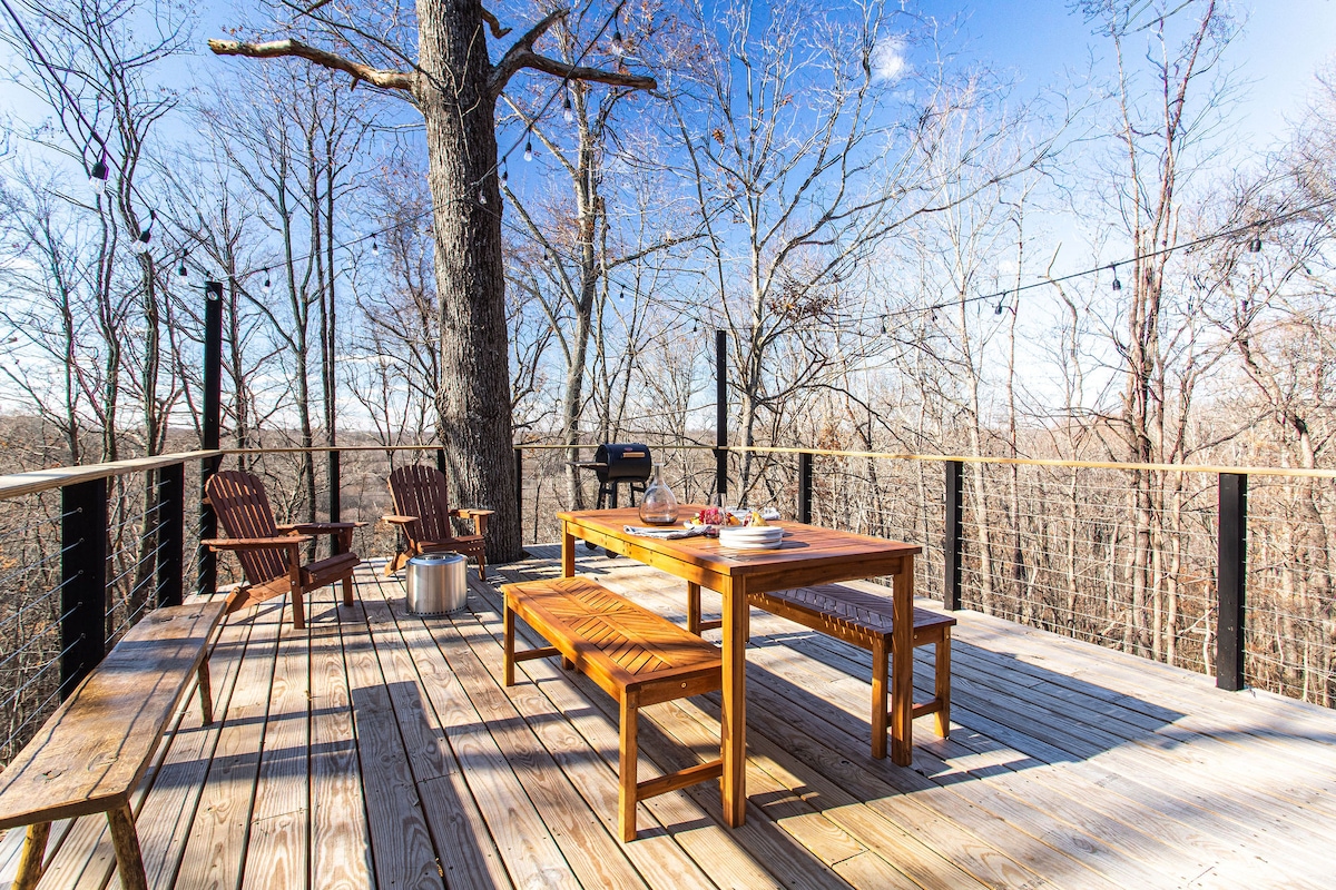 An outdoor deck is framed by trees, offering a spacious wooden dining table and bench seating. Two Adirondack chairs provide a cozy spot for relaxation. A grill is positioned nearby, with string lights overhead illuminating the space, creating a serene atmosphere in nature.