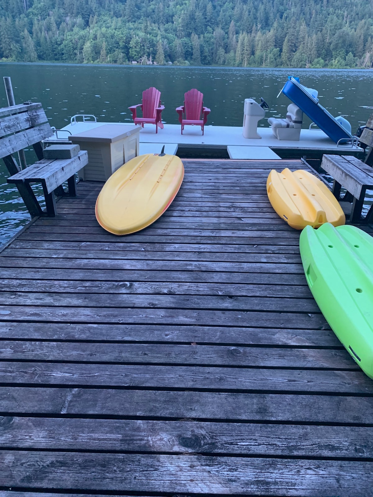 A wooden dock extends over the water, featuring two adirondack chairs in red. Two kayaks are positioned on the dock, one in yellow and one in green. The serene lake and surrounding greenery provide a tranquil backdrop.