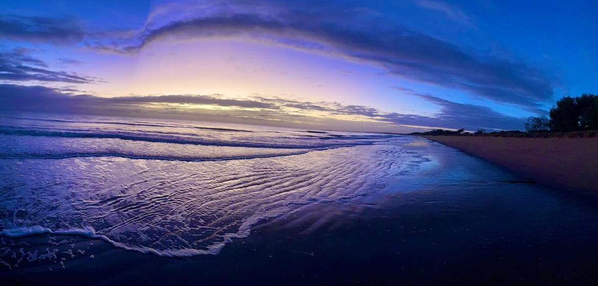 A panoramic view of the beach at twilight, showcasing gentle waves lapping the shore. The sky transitions from shades of purple to golden hues, reflecting on the wet sand. Sparse vegetation is visible in the distance, contributing to the serene coastal landscape.