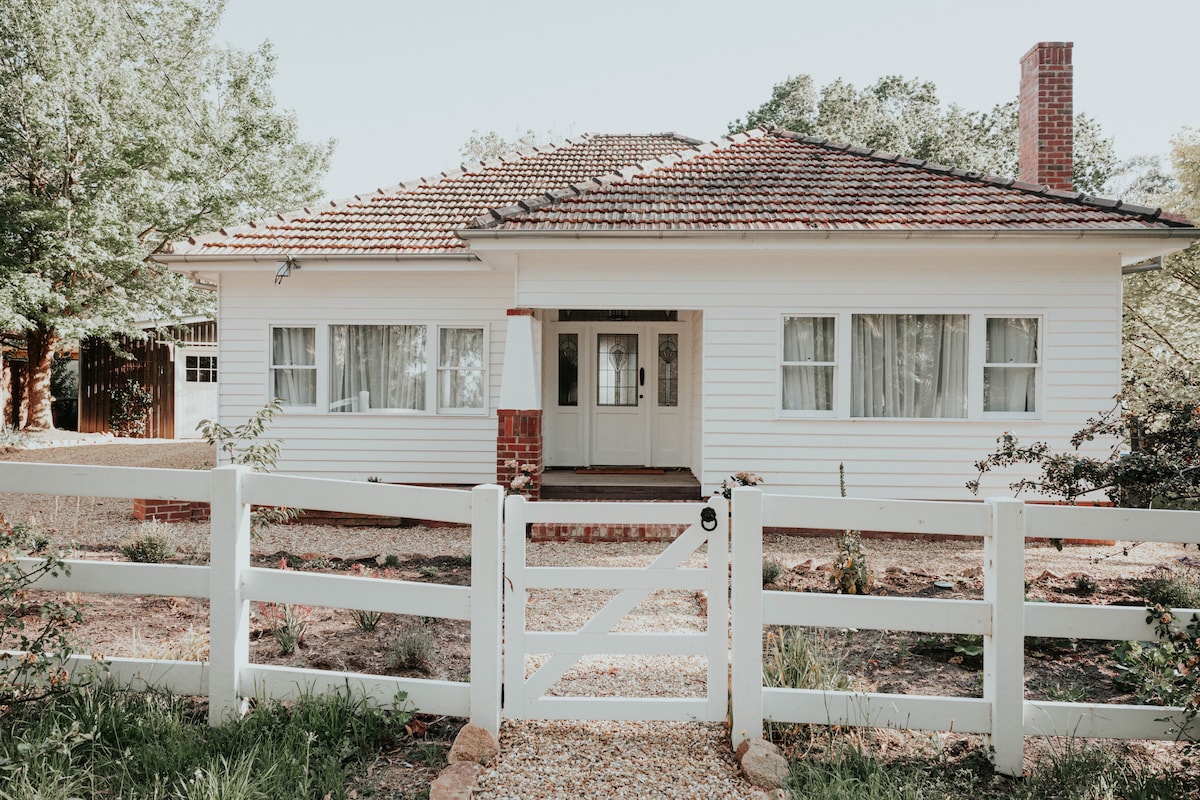 A charming cottage is framed by a white picket fence, featuring a covered front porch with double doors. Large windows allow natural light to fill the inviting entryway, while a well-kept garden area surrounds the home, showcasing its quaint and welcoming appearance.