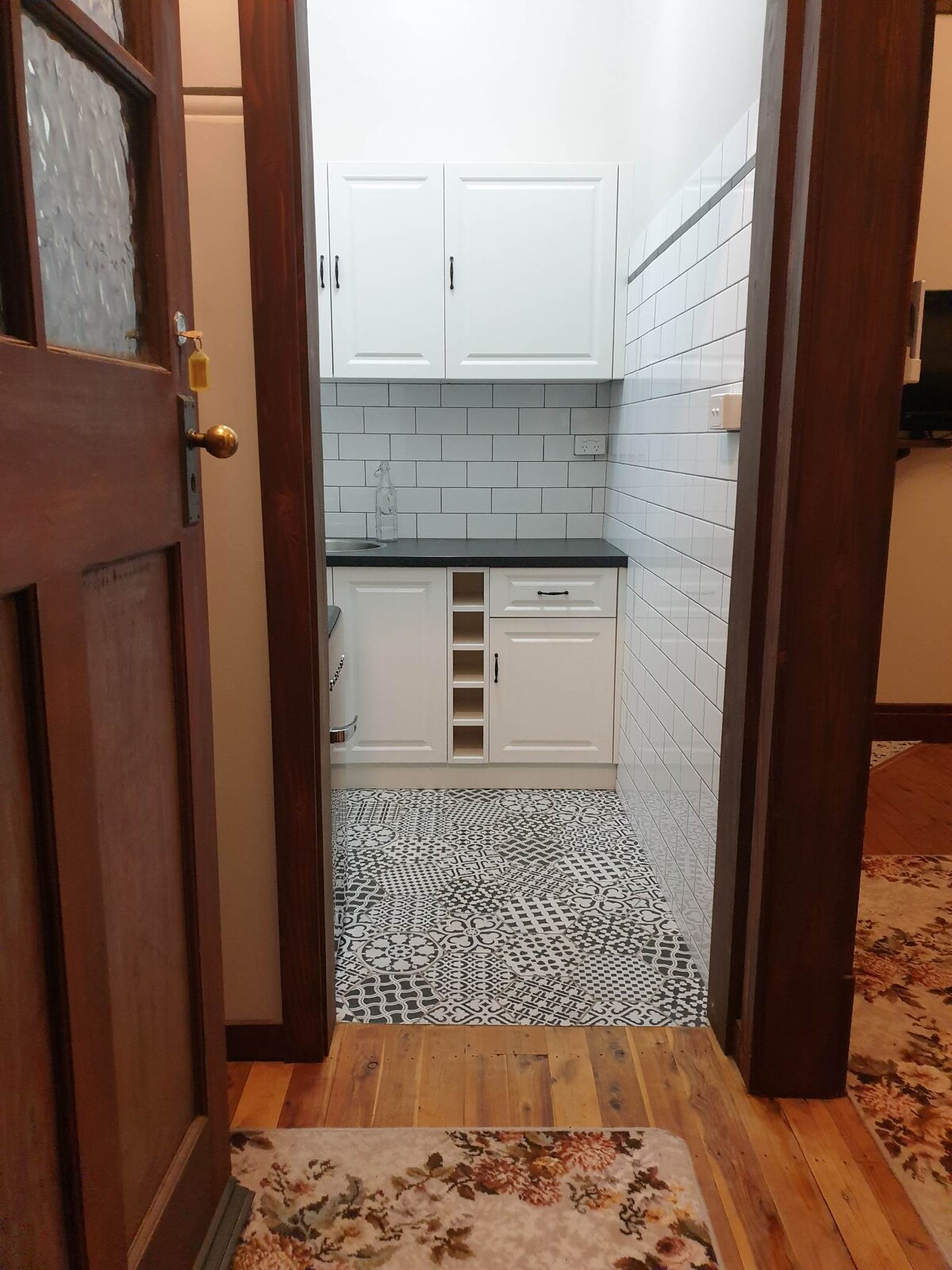 A compact kitchenette is visible, featuring white cabinets and black countertops. Decorative tiled flooring adds character, while a wine rack is positioned beneath a counter. The space is illuminated with natural light filtering in, enhancing the overall functionality of the area.
