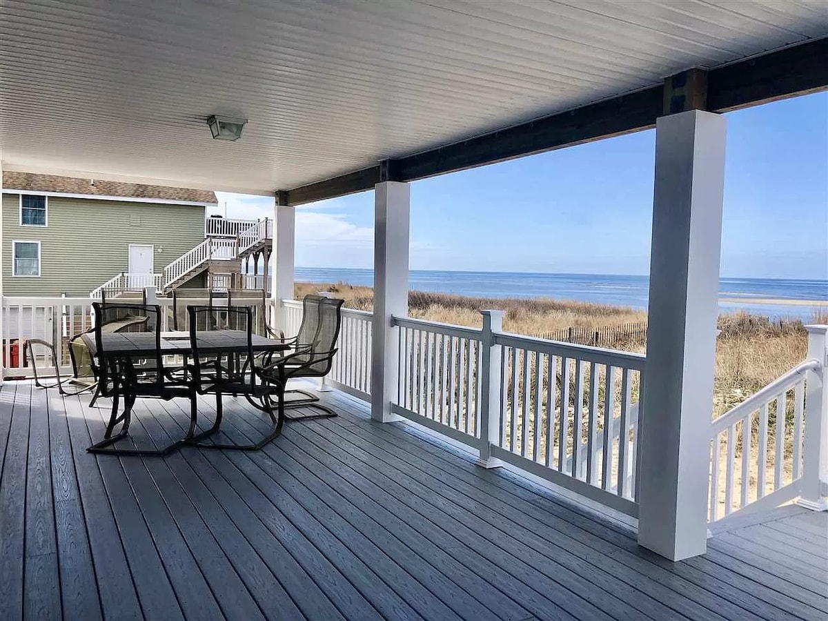 A covered outdoor patio is shown, featuring a dining table with six chairs. The views overlook the bay, with grassy dunes visible in the distance. The deck's wood flooring complements the white railing, and the bright sky enhances the serene setting.