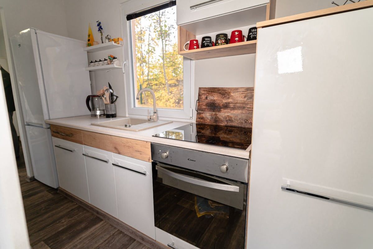A modern kitchen equipped with essential appliances, including a refrigerator and stove. White cabinetry is complemented by wooden accents. Natural light enters through a window framed by autumn foliage, enhancing the functional layout with ample counter space and organized storage above.