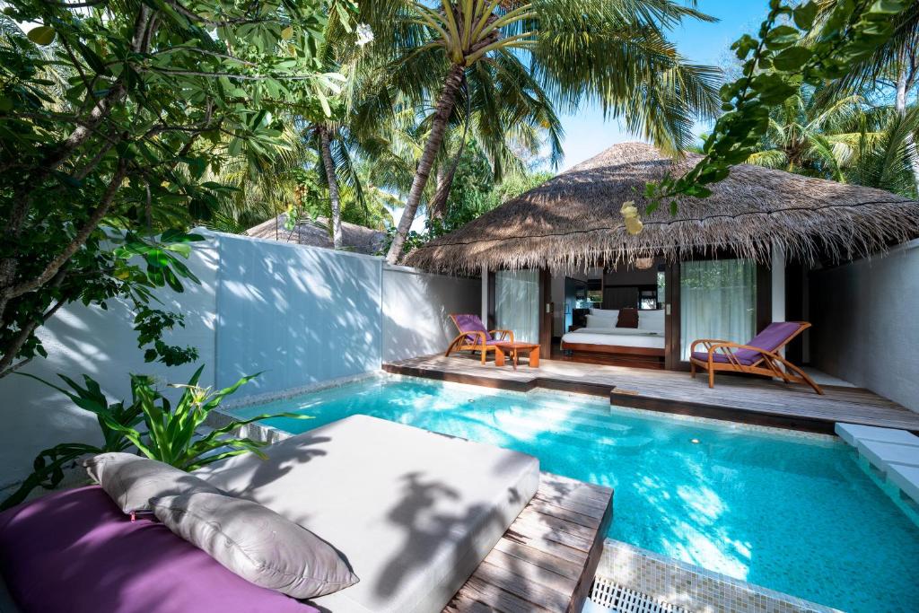A private pool surrounded by lush greenery is captured in this image. A thatched-roof structure is visible in the background, featuring large glass doors that lead to a cozy seating area. Sun loungers are positioned nearby, offering a tranquil space to relax.