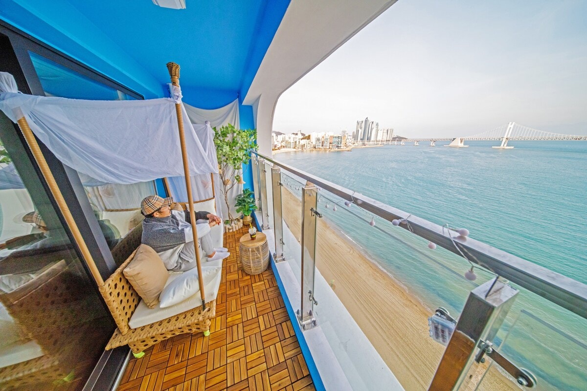 A terrace overlooking the ocean is presented, featuring a cozy seating area with a covered chair and a small potted plant. The water glistens under sunlight, while the distant city skyline and bridge create a serene coastal view.