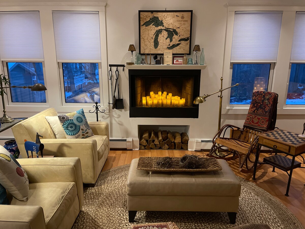 A cozy living area features a combination of light-colored seating and a wooden rocking chair. A decorative coffee table sits atop a textured rug. A fireplace is highlighted, surrounded by candlesticks and stacked logs, with windows allowing natural light to fill the space.