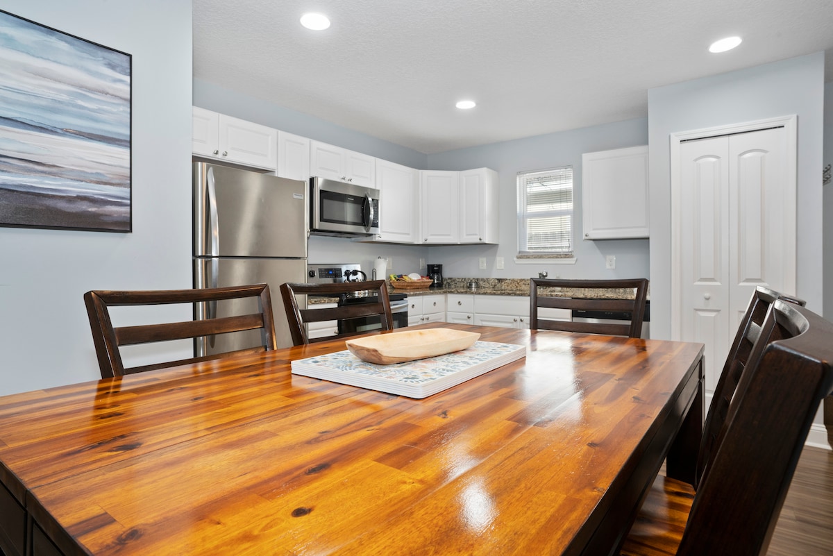 A spacious kitchen area is featured, with white cabinetry and stainless steel appliances. A wooden dining table, accommodating multiple seats, is positioned in the center. Natural light is provided through the adjacent window, highlighting the granite countertops and a piece of art on the wall.