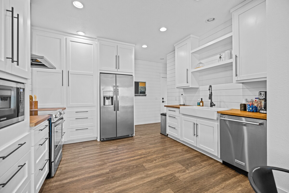 A modern kitchen features white cabinetry with a natural wood countertop. Stainless steel appliances, including a refrigerator and oven, are included. A large farmhouse sink is positioned beneath a window. The space is illuminated by recessed lighting and has a warm wood floor.