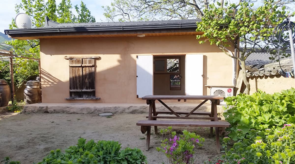 A rustic exterior of the accommodation is presented, featuring warm earthy walls and wooden shutters. A picnic table is situated in the garden, surrounded by greenery and colorful flowers. Natural light is observed illuminating the space, with an air conditioning unit visible on the side.