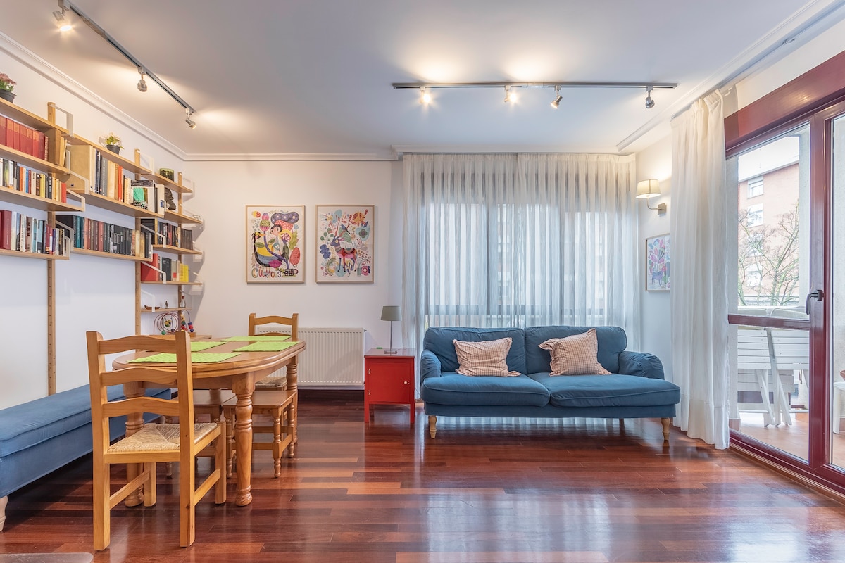 The living area features a blue sofa and a dining table surrounded by wooden chairs. A bookshelf is visible on the wall, and natural light filters through sheer curtains. A balcony can be seen in the background, enhancing the room's spaciousness.