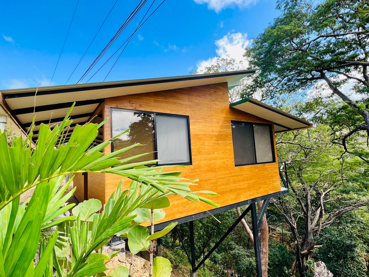 The cabin is elevated and constructed with natural materials, showcasing a modern exterior. Large windows allow for views of the surrounding trees and sky. The structure is supported by stilts, providing a unique vantage point that blends with the natural landscape.