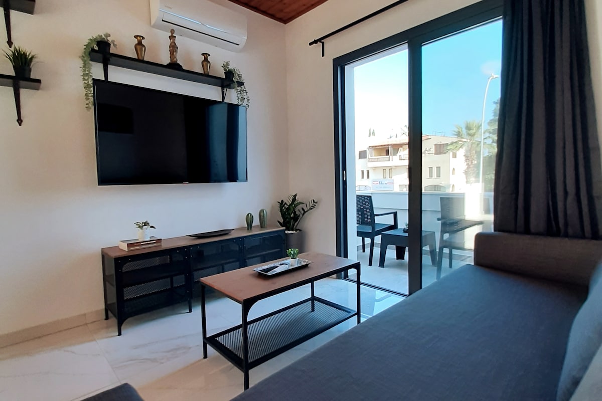 A modern living area features a large flat-screen TV mounted on the wall above a stylish media console. A comfortable sofa provides seating, and a coffee table is positioned in front. Floor-to-ceiling glass doors lead to a balcony, allowing natural light to brighten the space.