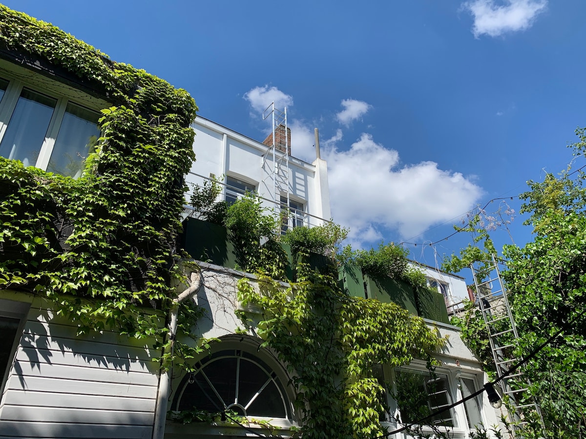 The building features a lush green facade, with climbing vines covering parts of the exterior. A scaffold indicates ongoing maintenance, while a clear blue sky and fluffy clouds provide a bright backdrop to this charming urban setting.