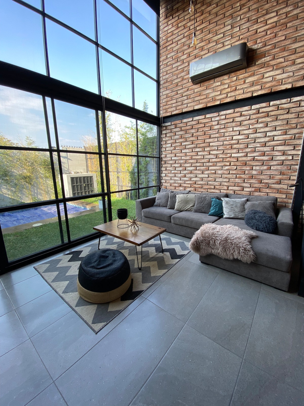 A stylish living area features large glass windows that frame the outdoor view. A gray sectional couch is complemented by decorative pillows, while a low coffee table sits on a patterned rug. A soft pouf adds an additional seating option, and sunlight fills the space.