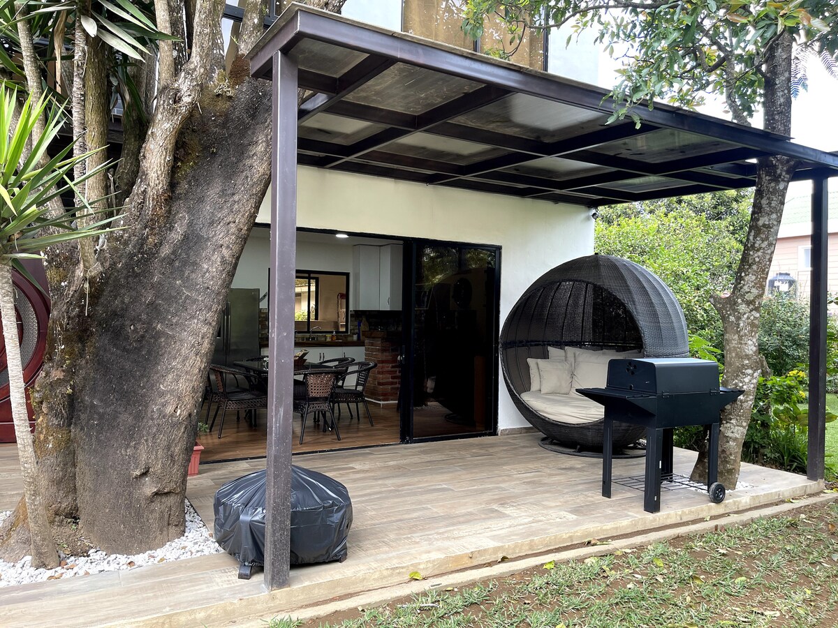 An outdoor patio area is sheltered by a sleek canopy, featuring a comfortable round seating pod and a barbecue grill. Lush greenery surrounds the space, while a wooden deck provides a natural contrast to the modern structure.