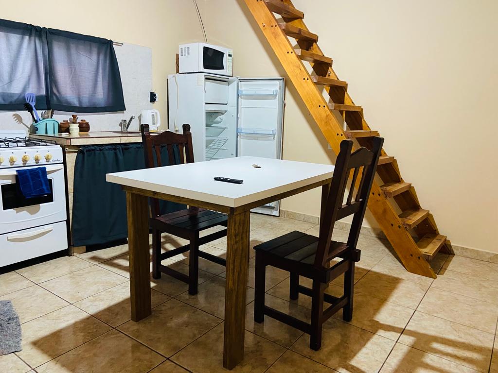 A dining area is furnished with a rectangular white table and two wooden chairs. A nearby kitchenette features a stove, microwave, and refrigerator. Wooden stairs ascend to a loft area, enhancing the space's functionality.