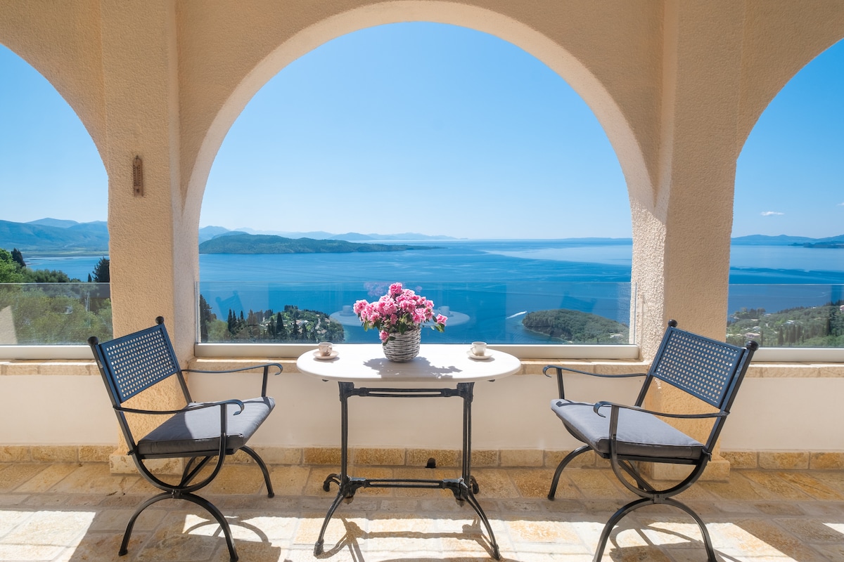 A terrace area is featured, showcasing two black metal chairs positioned around a small table. A vase of pink flowers adds a touch of color, while panoramic views of the sea and distant hills are framed by archways. Sunlight bathes the space, enhancing the tranquil setting.