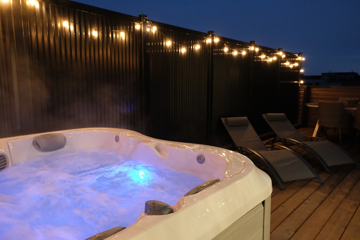 A private rooftop terrace is highlighted by a bubbling jacuzzi hot tub, featuring soothing blue lighting. The area is accented with string lights overhead, creating a cozy ambiance. Two lounge chairs and an outdoor dining area complete the space, inviting relaxation and enjoyment.