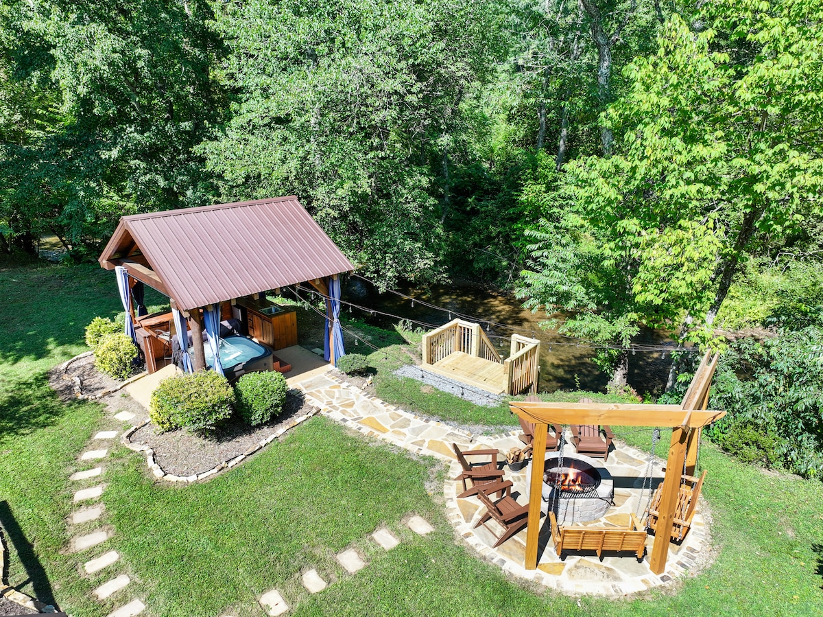 An outdoor area includes a covered pavilion with comfortable seating and a hot tub, surrounded by lush greenery. A stone pathway leads to an oversized firepit, featuring multiple Adirondack chairs, creating an inviting space for relaxation by Fightingtown creek.