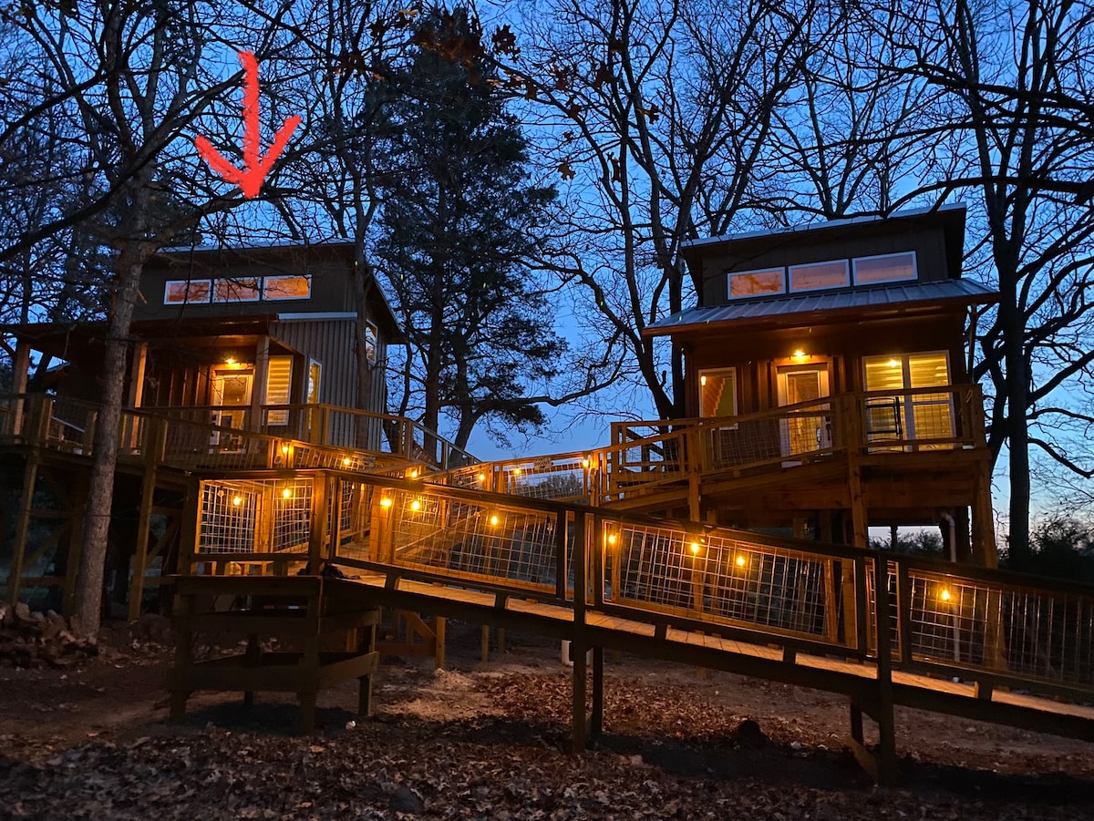 Two rustic treehouses are illuminated in the evening light, showcasing warm outdoor lighting against the darkening sky. Wooden walkways with railings connect the structures, surrounded by tall trees, creating a serene and inviting atmosphere.