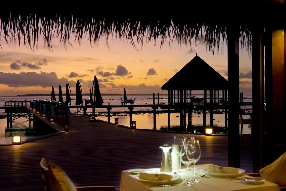 An outdoor dining setup is presented against a sunset backdrop. A table with elegant tableware is visible in the foreground. In the distance, a wooden pier extends into calm waters, flanked by lounge chairs and umbrellas, while the sky showcases a blend of warm hues.