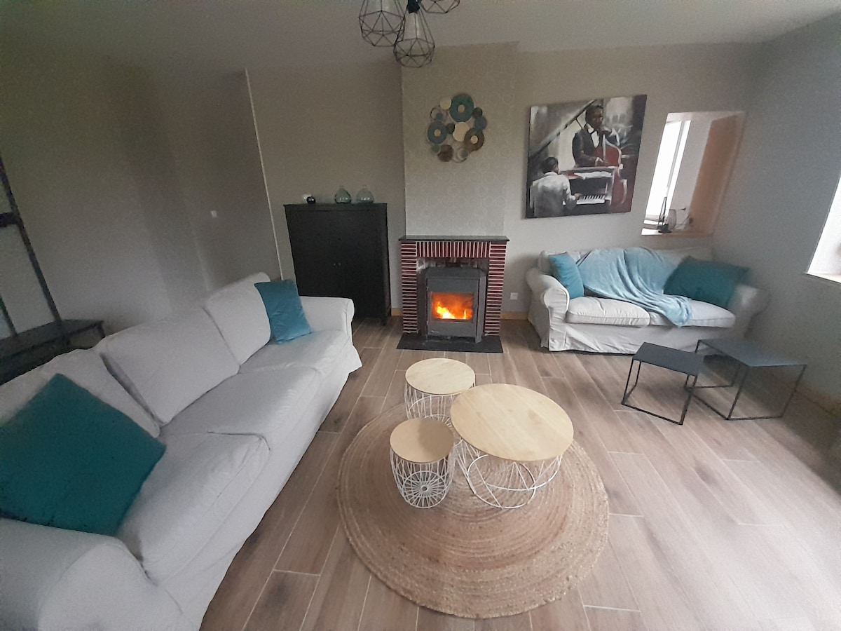 A spacious living area features two sofas, one draped with a soft blanket, alongside a modern fireplace emitting a warm glow. Two nested coffee tables are positioned on a circular woven rug, enhancing the relaxed ambiance. Natural light streams through the nearby window.