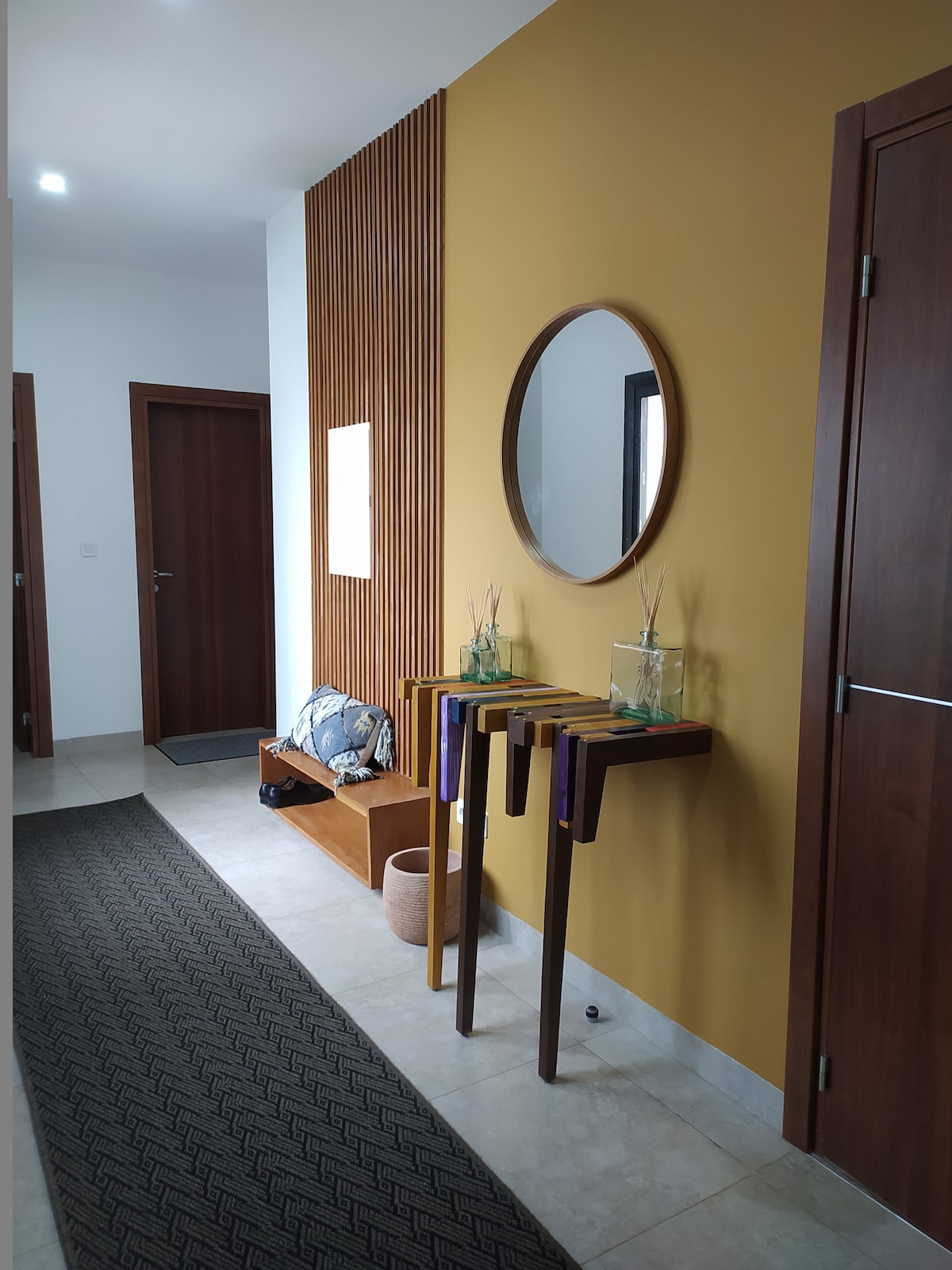 A modern entryway features a warm yellow accent wall and stylish wooden paneling. Two slender tables are positioned against the wall, each topped with decorative plants in glass vases. A round mirror reflects the space, while a bench and a textured rug enhance the welcoming feel.